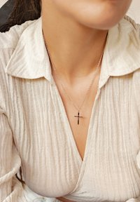 Collar de plata con colgante de cruz sobre una delicada cadena, llevado sobre una blusa texturizada de color beige claro.