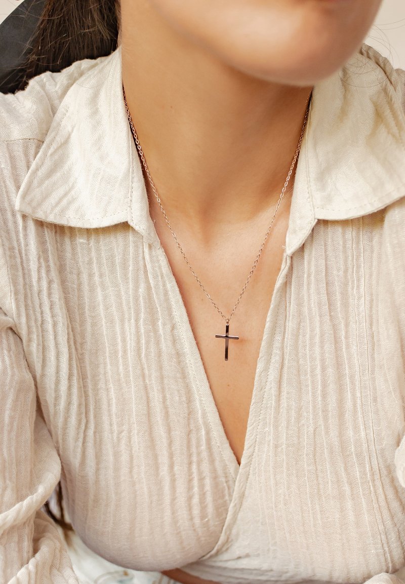Collar de plata con colgante de cruz sobre una delicada cadena, llevado sobre una blusa texturizada de color beige claro.