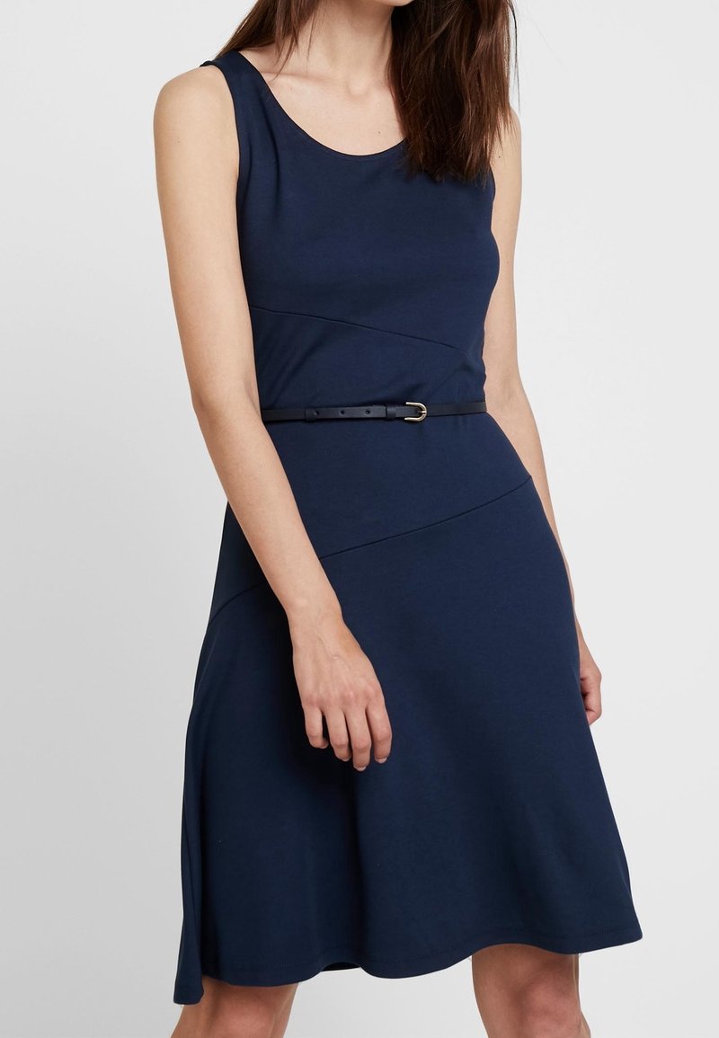 Femme portant une robe bleu marine sans manches, longueur genou, avec une fine ceinture noire, debout devant un fond gris clair uni.