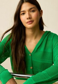 Grön stickad cardigan med V-ringning, utrustad med horisontella ränder och träknappar. Texturen är lätt och ventilerande.