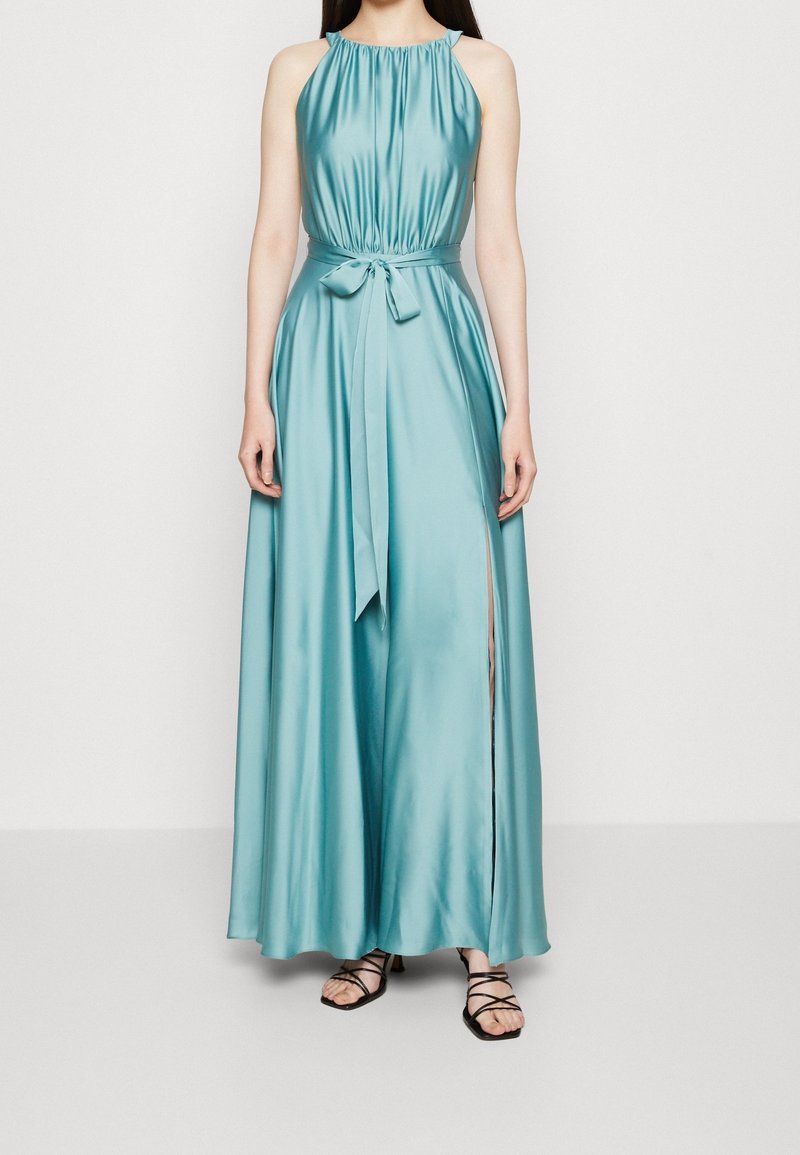 Femme portant une robe maxi sans manches en satin bleu clair avec une ceinture nouée à la taille et une fente haute sur un côté, associée à des sandales noires à fines lanières.