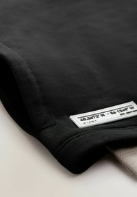 Sort bomulds T-shirt med ribbet tekstur og et hvidt rektangulært label med koordinater, foldet ved kanten.