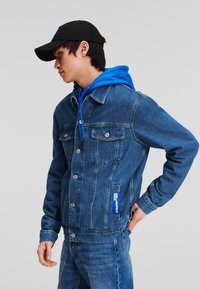 Jachetă din denim de culoare bleu închis, cu nasturi argintii, buzunare laterale și o etichetă de brand; purtată peste un hanorac albastru strălucitor și asortată cu blugi.