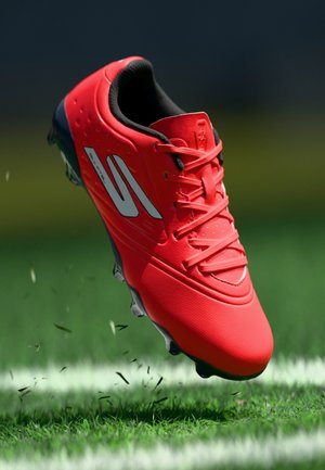 Spilgti sarkana futbola kurpes ar melnu zolīti un baltu logotipu, gaisā virs zāles laukuma pie baltas līnijas, ar apkārt lidojošiem zāles izcirtumiem.