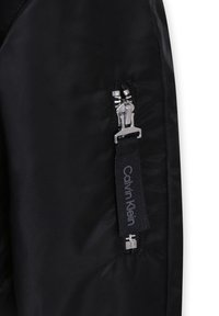 Giacca nera in nylon con zip argentata e etichetta con logo Calvin Klein sulla tasca. La texture è liscia con una finitura opaca.