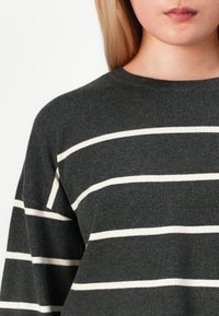 Blonde Person, der einen dunkelgrauen Pullover mit weißen horizontalen Streifen trägt, von der Brust bis zu den Lippen vor einem weißen Hintergrund gezeigt.