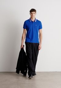 Kék póló csíkos gallérral, fekete nadrág, fekete kabátot tart, világos színű padlón sétál, sima fehér háttér előtt.