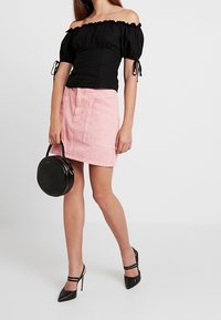 Blusa nera off-the-shoulder con texture arricciata, abbinata a una gonna di jeans rosa chiaro con tasche frontali, e tacchi neri con cinturini.