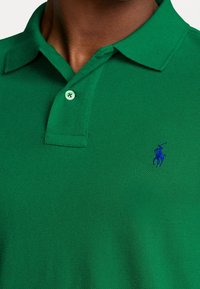 Grön pikétröja i strukturerad tyg, med klassisk krage, två knappar och en blå broderad logotyp på vänster bröst.