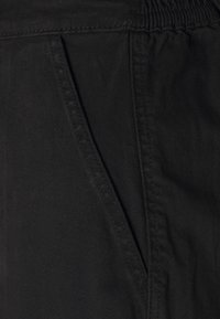 Gros plan d'un pantalon en tissu noir montrant une poche latérale avec des coutures visibles et une section de ceinture élastique.