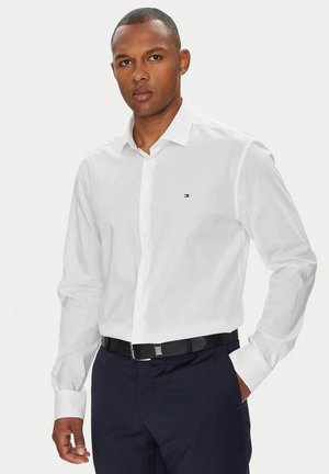 Uomo che indossa una camicia bianca con bottoni e un piccolo logo sul petto, pantaloni blu navy e cintura nera, in piedi con una mano in tasca.