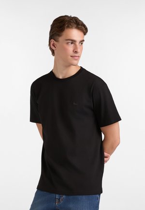 T-shirt nera in cotone, con colletto girocollo e maniche corte. Dettaglio del logo discreto sul lato sinistro del petto. Jeans visibili sotto.