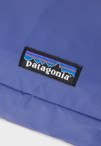 Borsa Patagonia con tessuto di un profondo colore viola, texture liscia e un'etichetta nera con grafiche colorate di montagne e testo bianco.