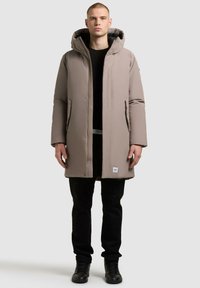 Parka imbottita beige con cappuccio, dotata di zip frontale a tutta lunghezza, tasche laterali e un'etichetta con logo all'orlo; abbinata a pantaloni neri e stivali.
