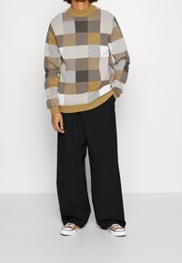 Sweter w kratę w kolorze brązowym, beżowym i białym. Długie rękawy, okrągły dekolt, ściągacze na mankietach i u dołu. Połączony z szerokimi czarnymi spodniami i brązowymi butami.