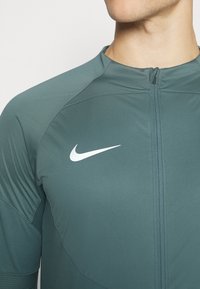 Felpa con zip Nike di colore verde scuro, caratterizzata da un design aderente, tessuto morbido, logo bianco sul lato sinistro del petto e sezioni delle spalle testurizzate.