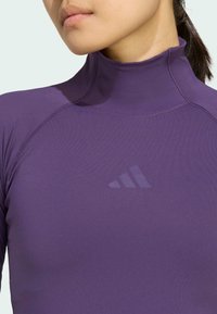 Haut à manches longues violet avec un col montant, arborant un logo Adidas discret. Fabriqué en matière lisse et extensible avec des coutures plates.