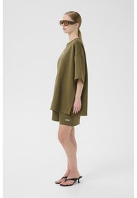 Olijfgroene oversized t-shirt en bijpassende shorts van zachte stof, met een zijsplit en geborduurd logo. Gecombineerd met bruine zonnebril.