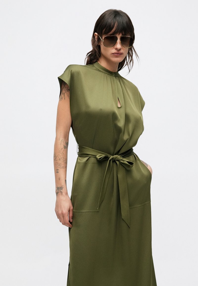 Olive green dress with a high neck, short sleeves, and a keyhole detail. Features a matching tie belt and side pockets for functionality.