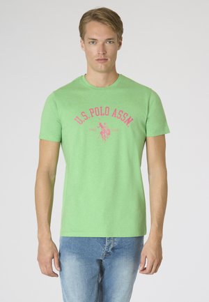 Giovane uomo che indossa una maglietta verde chiaro U.S. Polo Assn. con logo rosa e jeans azzurri, in piedi davanti a uno sfondo bianco semplice.