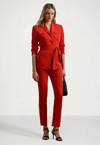 Sieviete valkā sarkanu jostotu bleizeri ar saskaņotiem biksēm un melnā augstpapēžu sandales, rokās tur mazu melnu rokassomu.