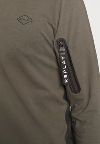 Olivgrünes Langarmshirt mit einem Diamantlogo auf der Brust und einer Reißverschlusstasche auf dem linken Ärmel, mit schwarzen Akzenten.
