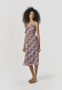 Ärmelloses Kleid mit floralem Muster aus leichtem Stoff, mit Rüschen am Ausschnitt, geraffter Taille und mittellang bis zur Wade. Kombination mit flachen Sandalen.
