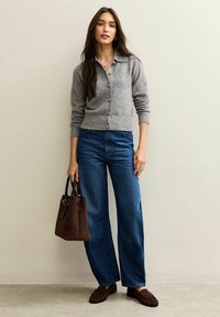 Grå kofta med knäppning framtill och ribbade muddar; blå vida jeans; brun texturerad handväska; bruna loafers. Neutral bakgrund.