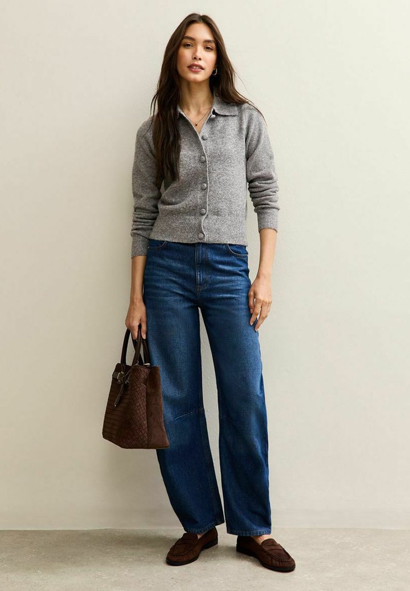 Grå kofta med knäppning framtill och ribbade muddar; blå vida jeans; brun texturerad handväska; bruna loafers. Neutral bakgrund.