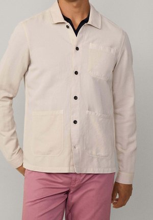 Mann trägt eine hellbeige Jacke mit Knöpfen und drei Fronttaschen über einem schwarzen Hemd und einer rosa Hose, steht vor einem schlichten Hintergrund.