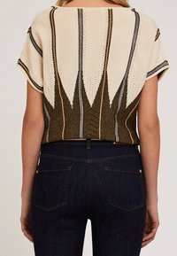 Beige gestricktes Oberteil mit grünen und metallischen Streifen. Es verfügt über ein geometrisches Muster, kurze Ärmel und einen lockeren Schnitt, kombiniert mit dunkelblauen Jeans.