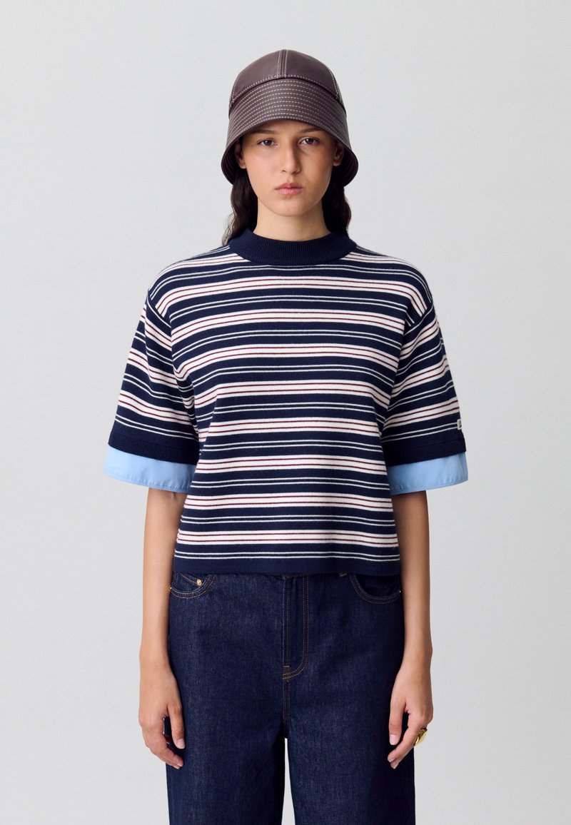 Jersey de rayas en azul marino y crema con mangas cortas, con puños en azul claro. Combinado con pantalones de mezclilla oscuros.
