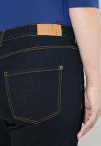 Tmavě modré džíny s hladkým povrchem, žlutým šitím a hnědým koženým značkovým štítkem na pásku. Obsahují dvě zadní kapsy.