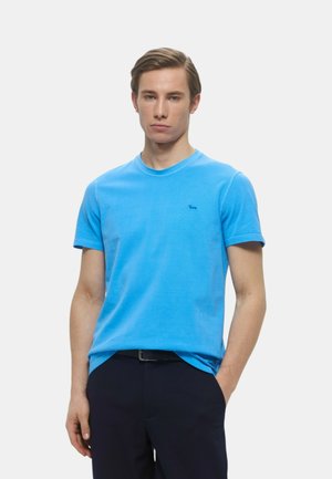 T-shirt di cotone azzurro chiaro con scollo rotondo e maniche corte. Presenta un piccolo logo sul lato sinistro del petto. Indossato con pantaloni scuri.