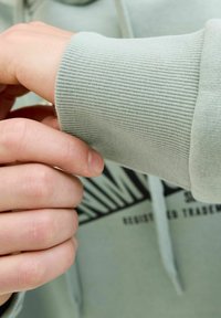 Haut à capuche vert clair avec des poignets côtelés. Gros plan sur une main saisissant le poignet, montrant la texture lisse et le détail du logo sur le tissu.