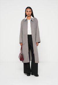 Hosszú szürke gyapjúkabát gallérral, két első zsebbel és oldalsó cipzárokkal. Fehér inggel és fekete nadrággal viselve, kiegészítve bordó táskával.