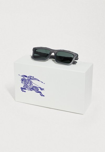 Γκρι γυαλιά ηλίου με σκούρους φακούς, τοποθετημένα πάνω σε ένα λευκό κουτί με εκτυπωμένο μπλε λογότυπο αλόγου. Το κουτί έχει λεία υφή και ορθογώνιο σχήμα.
