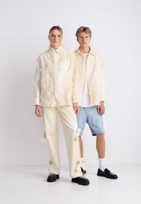 Μπεζ υφασμάτινο σακάκι και εκρού παντελόνια με φιόγκους στο τελείωμα, σε συνδυασμό με τζιν σορτς και μαύρα παπούτσια χωρίς κορδόνια, πάνω σε λευκό φόντο.