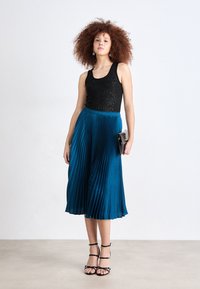 Top senza maniche nera e scintillante abbinato a una gonna midi plissettata color teal. La modella indossa sandali neri con cinturini e tiene una piccola clutch nera.
