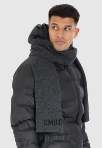 Grau gestrickter Schal, der um den Hals drapiert ist, mit einem strukturierten Stoff und dem Wort "SMILO" in Schwarz am Ende, kombiniert mit einer schwarzen Daunenjacke.