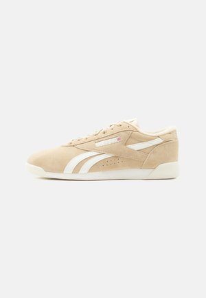 Beige suède lage sneaker met witte Reebok-strepen, veters en een gedempte zool, van opzij gezien tegen een witte achtergrond.