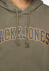 Oliwkowa zielona bluza z kapturem z nadrukiem na przodzie "PACK & JONES" w niebieskim i pomarańczowym kolorze. Posiada sznurki z frędzlami oraz miękką fakturę.