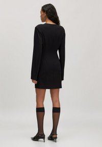 Vrouw die wegkijkt in een strak zwart langslipaansluitend mini-jurk, doorschijnende kniekousen en zwarte kitten heels.