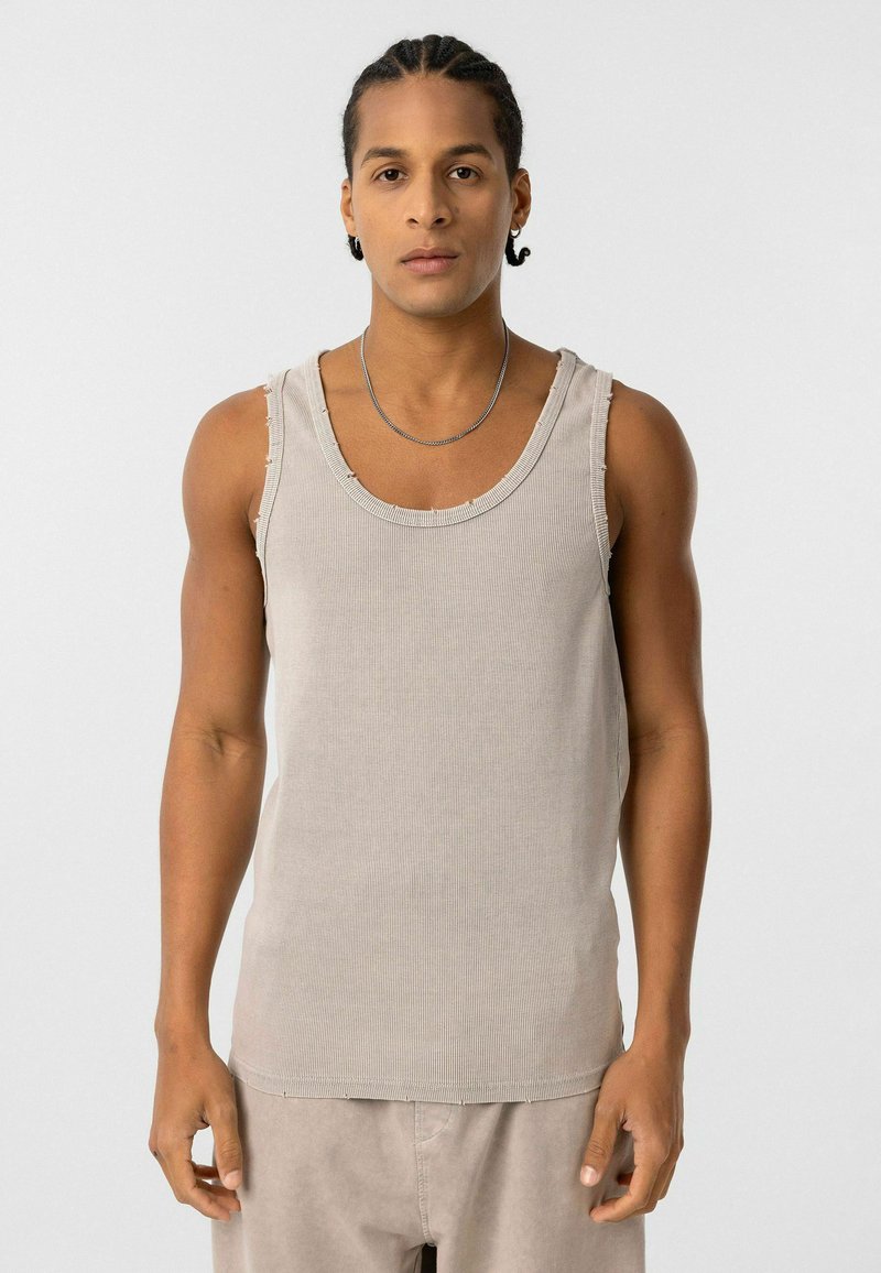 Beige geribbelde tanktop met een ronde halslijn en versleten randen, gemaakt van zachte katoenen stof. Model draagt het met lichtgekleurde broeken.