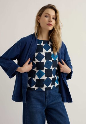 Mujer con blusa de estampado azul, chaqueta azul marino y jeans de mezclilla posando frente a un fondo liso.