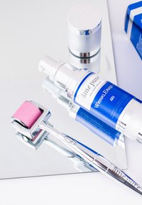 Blaue und silberne Hautpflegeprodukte auf einer reflektierenden Oberfläche: eine Gelpumpflasche, ein silbernes Rollgerät mit einem rosa Kopf und ein silberner Stift.