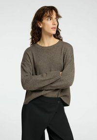 Maglione marrone a coste con scollo rotondo e vestibilità morbida, abbinato a pantaloni neri con dettaglio dei bottoni laterali.