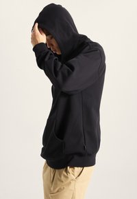 Felpa nera con cappuccio realizzata in tessuto morbido, dotata di tasca frontale a marsupio e cappuccio con cordino. Vestibilità classica con polsini e orlo a coste.