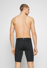 T-shirtlös man med kort blont hår vänd bort, iförd åtsittande svarta atletiska kompressionsshorts mot en enkel ljus bakgrund.