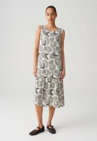Sleeveless midi dress in cream with black geometric wave pattern, featuring a ruffled hem. Worn with black mesh flats. Smooth fabric texture.
